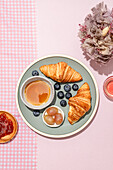 Top view of delicious croissants served on ceramic plate with fresh blueberries and jam placed on pink table