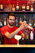 Focused bartender preparing alcohol cocktail and mixing ingredients in shaker while standing at counter in bar and looking at camera