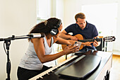 Fröhliche hispanische Frau mit lockigem Haar und Kopfhörern, die am elektronischen Klavier sitzt und einer lächelnden Freundin, die mit einer akustischen Gitarre in einem hellen Raum sitzt, ihr Smartphone zeigt