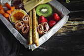 From above brunch box with assorted sliced meats various types of cheese and crispbreads arranged near ripe cup kiwi sweet strawberries and peeled mandarin near jam in glass jar on wooden table near napkin