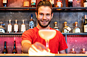 Cheerful barman serving glass with alcohol cocktail on counter in bar and looking at camera