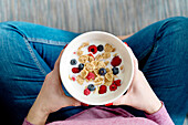Top view of crop unrecognizable female with bowl of cornflakes and ripe berries with milk for breakfast in light room