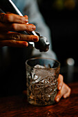 Crop female barkeeper putting ice cube with tongs into glass while preparing cocktail in bar with bottles on blurred background