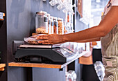 From above side view anonymous young woman pressing buttons on digital balance while weighing plastic bag with chocolate cornflakes during work in eco friendly grocery shop