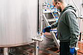 Seitenansicht eines männlichen Arbeiters mit Maske, der das Ventil eines Metalltanks bei der Arbeit in einer Brauerei während einer Pandemie wäscht