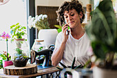 Smiling female florist with potted plant sitting in flower shop while speaking on mobile phone and getting order