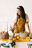Positive adult Hispanic female designer with long dark hair in apron smiling while decorating table with reed plants pumpkins and candles for Halloween celebration