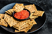 Red fresh trout fish caviar in a bowl served with sesame seed crackers on concrete table background