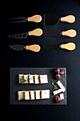 Draufsicht auf verschiedene Messer in der Nähe von leckerem Käse und Trauben auf schwarzem Brett