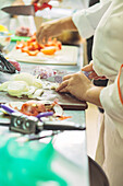 Side view of crop anonymous female cooks in uniform standing at counter cutting vegetables while preparing food in restaurant kitchen