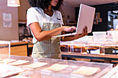 Cropped unrecognizable young African American woman in uniform and glasses with curly hair checking goods in glass boxes and using laptop while working in zero waste grocery store