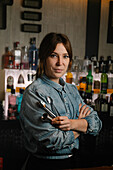 Attractive female bartender with jigger and tong in hands looking at camera while standing in bar with various alcohol drinks