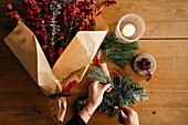 High angle of crop unrecognizable female florist standing and cutting twigs of fir while arranging Christmas bouquet on wooden table