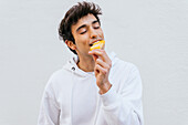 Happy young male with eyes closed eating sweet orange ice lolly enjoying taste on white background