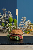 Fresh appetizing mini burger served on black board near blooming flowers against blue wall