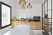 Moderne Kaffeemaschinen und Kaffeemühlen stehen auf einem Holztresen in einem hellen, modernen Kaffeehaus mit einem weißen Tisch am Fenster und Holzregalen
