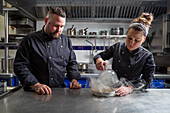 Male and female chefs in dark uniform removing dome lid from tasty dish with smoke while working in modern restaurant kitchen