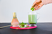 Crop anonymous female making healthy matcha tea with milk in glass mug placed near bamboo chasen and chashaku on table against white background