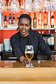 Cheerful black female barkeeper serving glass of refreshing beer in bar looking at camera