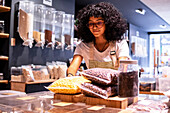 Positive young black female in uniform and glasses smiling and putting plastic bags with puffcorn snacks on shelf near jar of tea leaves during work in zero waste grocery store