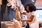 Cheerful young African American woman in casual clothes with curly hair smiling and using dispenser to fill paper bag while visiting eco friendly grocery shop