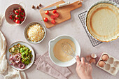 Top view of unrecognizable woman using whisk to beat eggs in bowl near fresh ingredients and crust while preparing quiche in kitchen at home