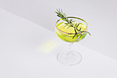 High angle of refreshing yellow alcohol cocktail garnished with fresh rosemary sprig placed on white table in studio