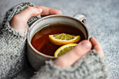 Kid hands in mittens hold cup of tea with lemon on grey concrete background