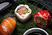 Tasty fresh assorted sushi served on green plant twigs on black plate with soy sauce on marble table near chopsticks