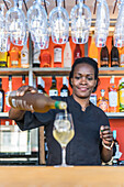 Focused African American female barkeeper pouring alcohol in glass with ice cube while making cocktail in bar