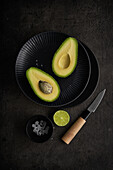 From above of fresh halves of avocado arranged on table with green lime, salt and knife on dark background
