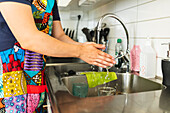 Crop unrecognizable person wearing apron and standing near kitchen sink filled with glasses with dish wash detergent placed on edge while washing hands in soft daylight