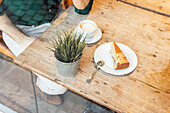 From above cropped unrecognizable male entrepreneur drinking hot coffee while sitting at wooden table with slice of cake in light modern cafeteria during lunch break
