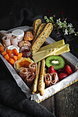 From above brunch box with assorted sliced meats various types of cheese and crispbreads arranged near ripe cup kiwi sweet strawberries and peeled mandarin near jam in glass jar on wooden table near napkin