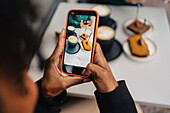 High angle of crop anonymous African American female blogger recording video of late coffee and cakes on cellphone at meeting with friend in outdoor cafe