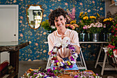 Cheerful female florist making flower wreath with ribbon bow while working in floral shop looking at camera