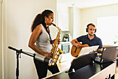 Hispanic Frau mit langen lockigen Haaren stehen und spielen Saxophon in der Nähe von positiven männlichen in Kopfhörer sitzen und spielen akustische Gitarre in hellen Raum in der Tageszeit
