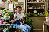 Smiling female florist with potted plant sitting in flower shop while speaking on mobile phone and getting order