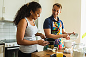 Happy couple in casual wear standing at high wooden kitchen table while mixing ingredients for bakery in bowls in daylight