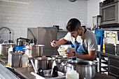 Focused young Hispanic male chef with dark hair in casual clothes and apron adding dry broth into saucepan while cooking in restaurant kitchen with modern appliances