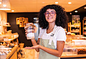 Happy young ethnic woman in uniform and glasses smiling holding glass jar with nuts and diverse flours to camera while working in zero waste grocery shop