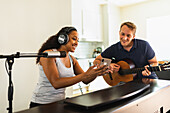 Fröhliche hispanische Frau mit lockigem Haar und Kopfhörern, die am elektronischen Klavier sitzt und einer lächelnden Freundin, die mit einer akustischen Gitarre in einem hellen Raum sitzt, ihr Smartphone zeigt