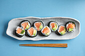 From above set of tasty Futomaki Norwegian sushi rolls with salmon and avocado served near chopsticks on blue background in light studio
