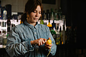 Focused female bartender taking off lemon zest while preparing for making cocktail in bar with various alcohol drinks during work