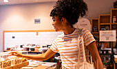 Side view of happy young African American woman in casual clothes with curly hair smiling and using tote bag while visiting eco friendly grocery shop