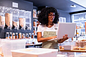 Young African American woman in uniform and glasses with curly hair checking goods in glass boxes and using laptop while working in zero waste grocery store