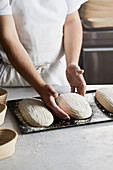 Unrecognizable person in apron removing loaves of sourdough bread from proofing baskets and arranging on cooking tray on table in kitchen