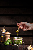 Crop anonymous person with spoon adding olive oil into pot with freshly prepared pesto sauce