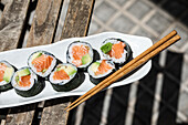 Draufsicht auf leckere japanische Futomaki-Sushi-Rollen aus norwegischem Lachs, die mit Stäbchen auf einem gepflasterten Gehweg in einer sonnigen Straße in der Stadt platziert werden