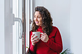 Zufriedene Frau mit einer Tasse Heißgetränk, die lächelnd aus dem Fenster schaut, während sie in einem hellen Raum zu Hause steht